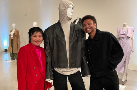 A man and woman posing with a male fashion mannequin wearing     black jeans and a black leather-like jacket over a light tan hooded garment