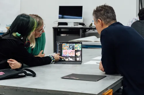 Three students hover over a laptop discussing a design showing on its screen