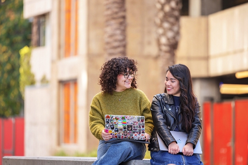 Two women students with laptops, sitting on a low outdoor wall on campus, talking to each other