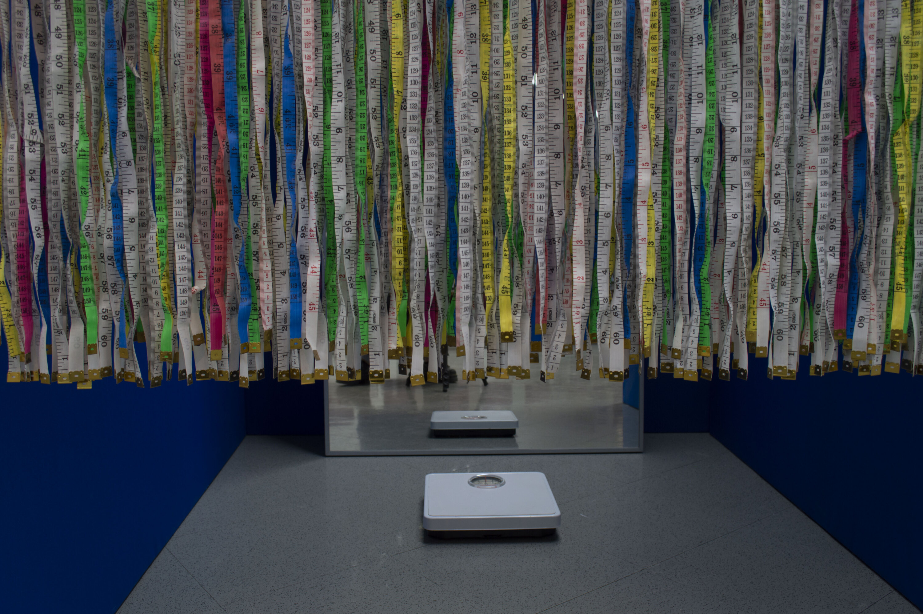 installation of measuring tapes hanging in front of a blue room. Inside room is a mirror and scale.