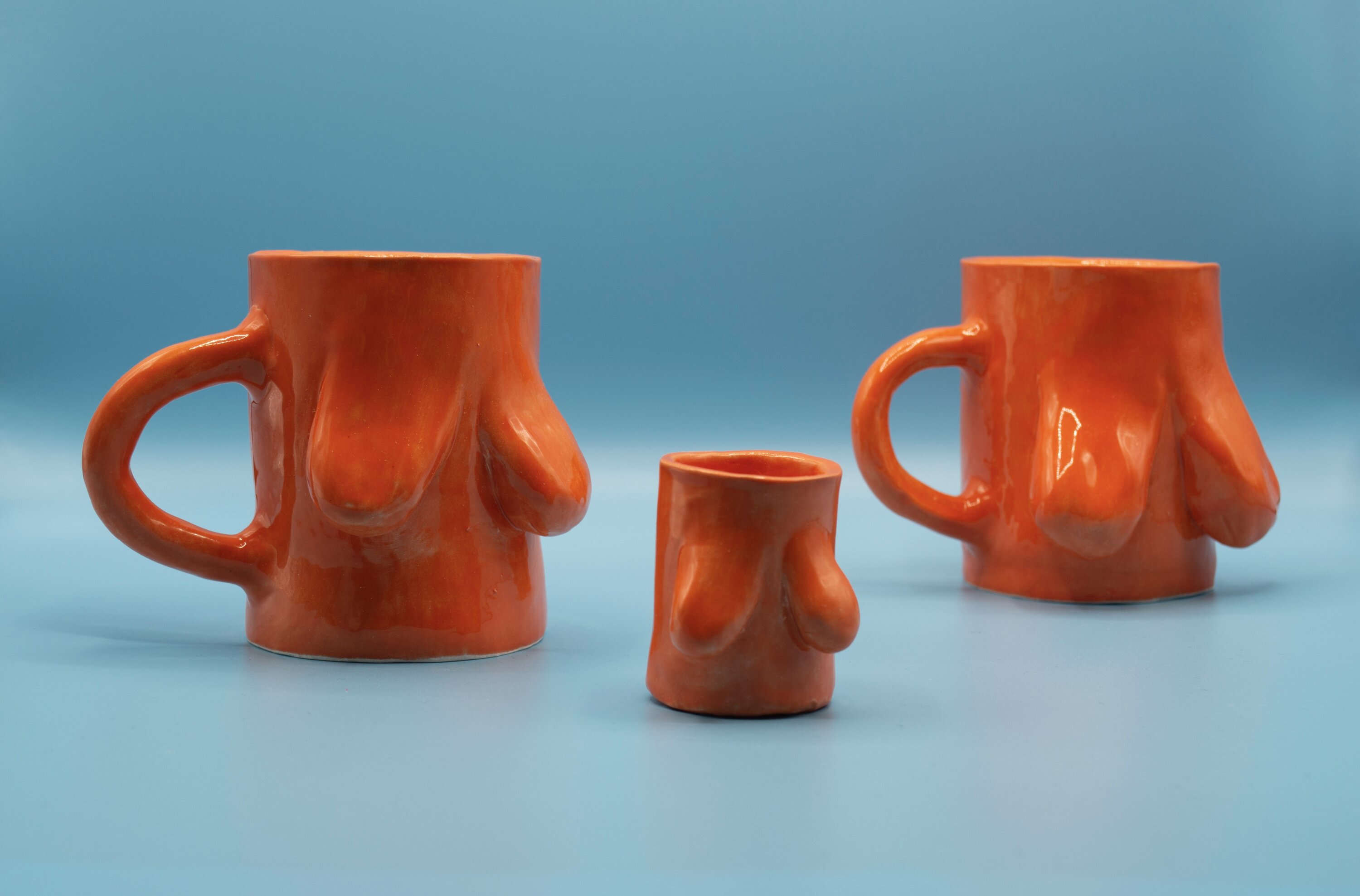 Two orange ceramic mugs and a shot glass in a blue background. Ceramics have breasts on them