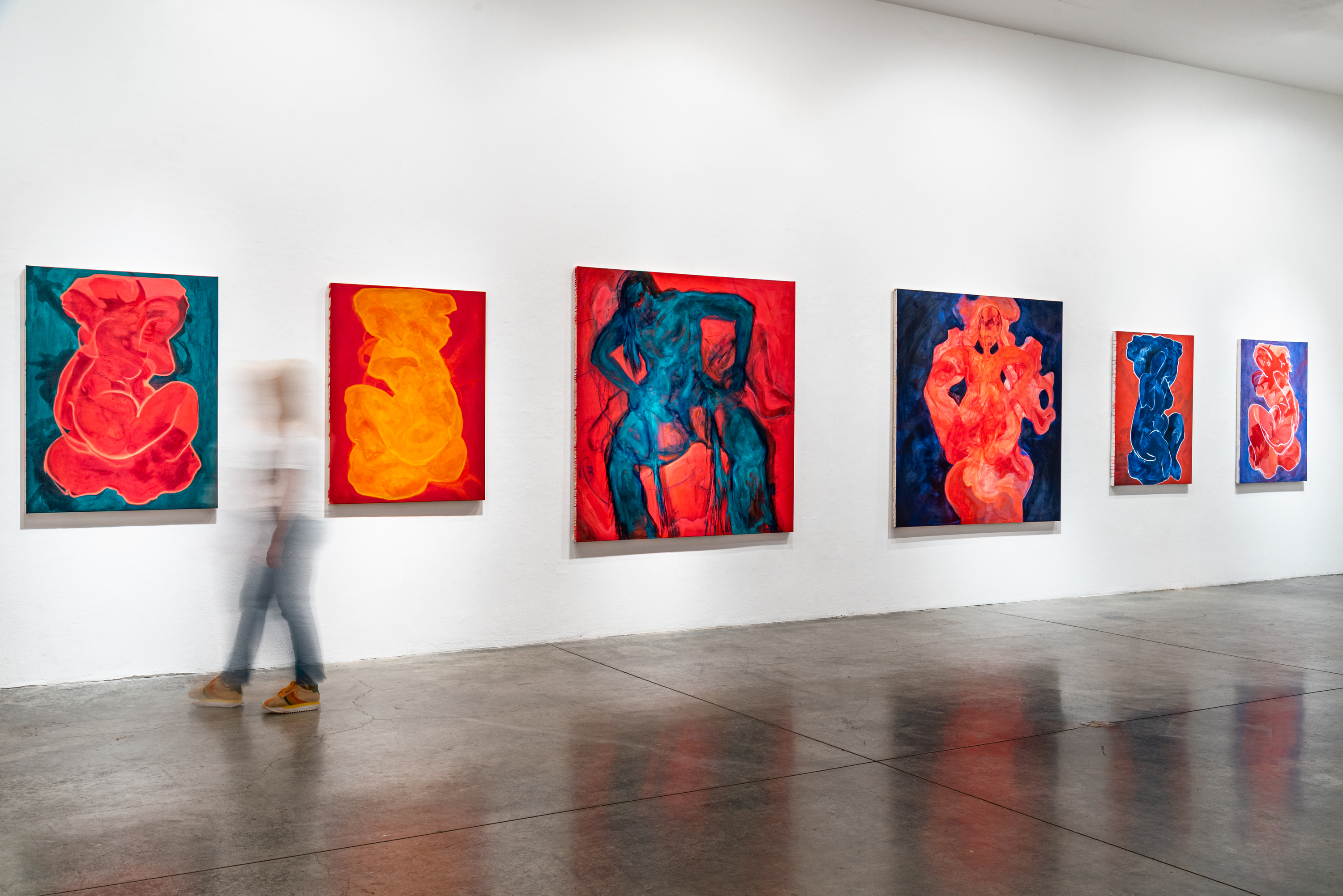 Installation view of a blurred figure walking past a wall of paintings