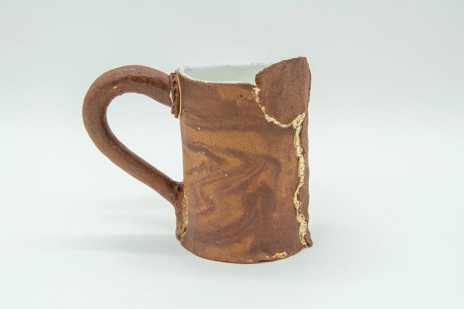 A handmade mug with an unglazed exterior, no effort was made to hide where the clay connects to itself. It leans slightly to the left towards the heavy handle. A red clay and a brown clay marble together while the seams of the mug are white with a third clay color, highlighting them.. The inside is glazed a matte white.