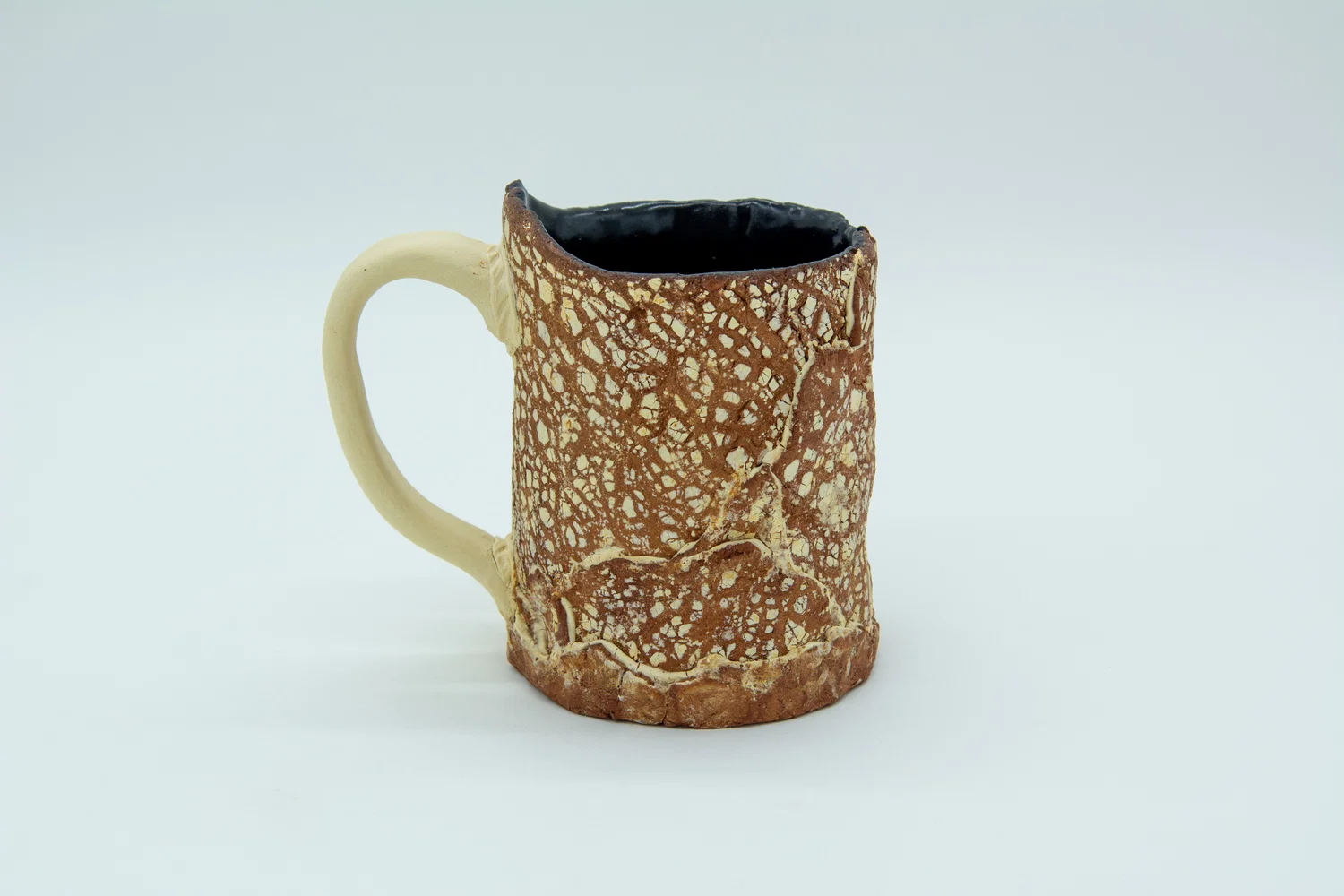 A handbuilt ceramic mug made of red and brown clay marbled together; It has a dry lake bed texture with the raised areas being a white clay. It’s made of different pieces of clay, making the marbling and texture inconsistent. White clay also fills the seams between the pieces, no effort was made to hide the seams. The interior of the mug is glazed black, and the handle is made of the same white clay that’s used in the seams.