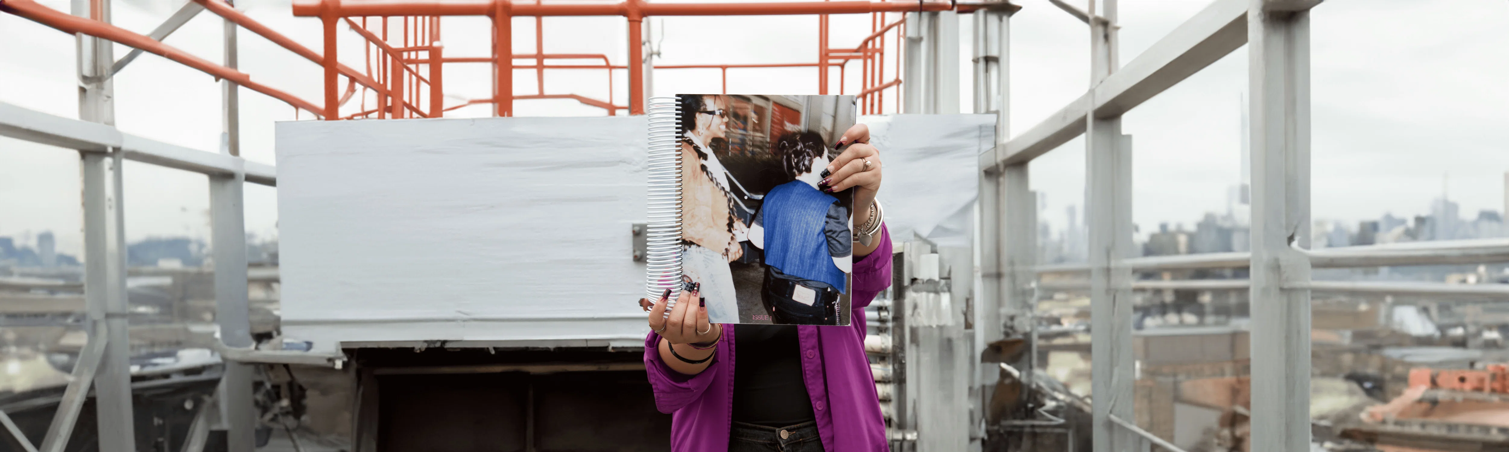 Aurora stands on an urban rooftop, holding up a spiral-bound copy of Sfitinzia, an editorial exploring fashion and subculture.