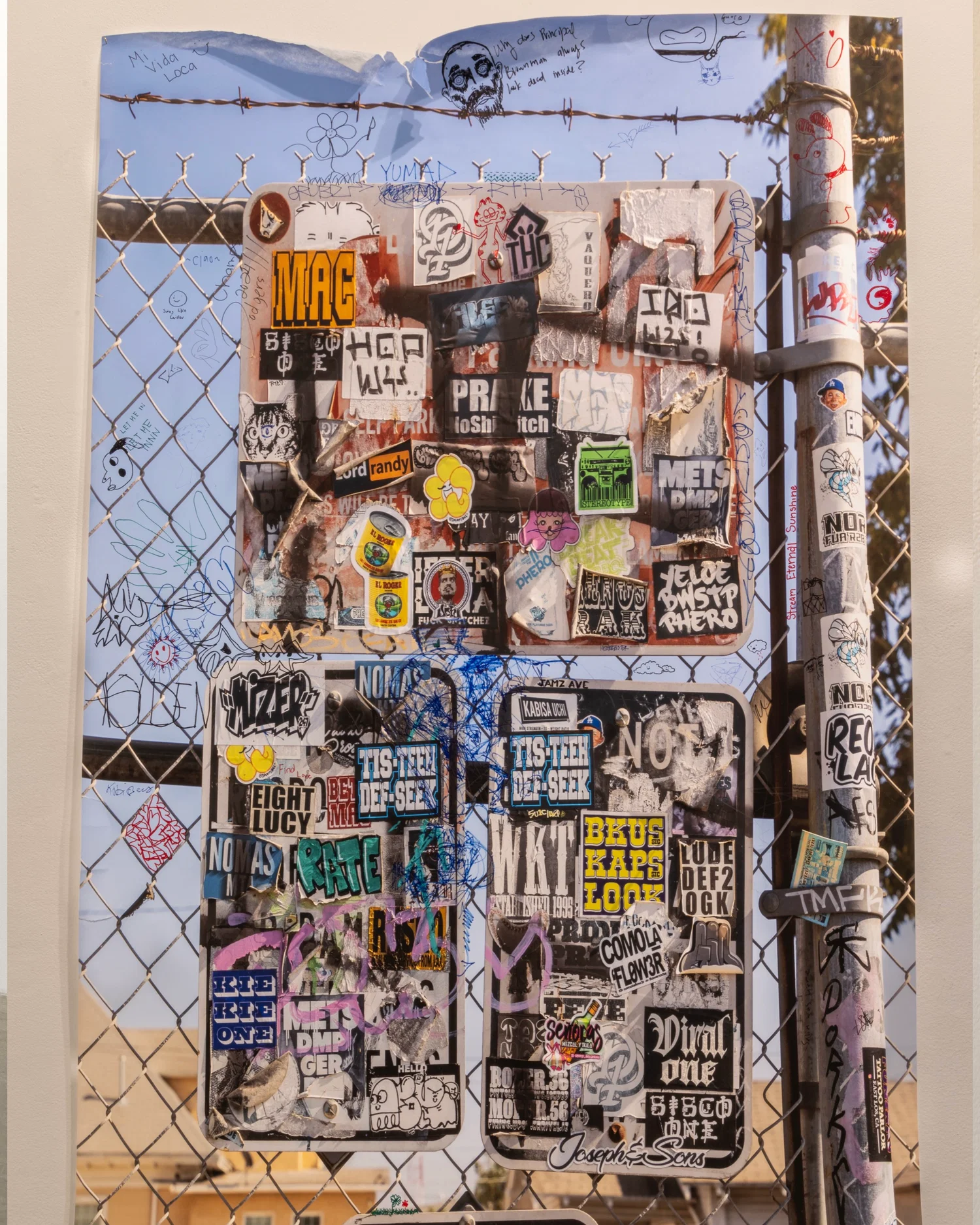 This image includes a print 60x82 image of a sign with stickers bombed all over in front of a community on a chain linked fence. On top of the print has various tagging and drawings
