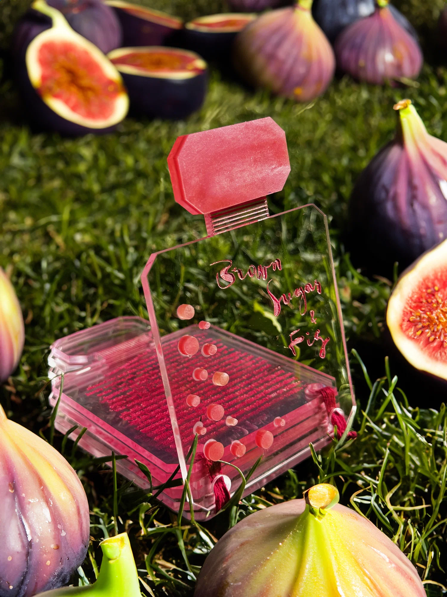 A small acrylic book in the shape of a perfume bottle, with the words "Brown Sugar and Fig" laser engraved on the front. The bottle is surrounded by images of fresh figs on grass.
