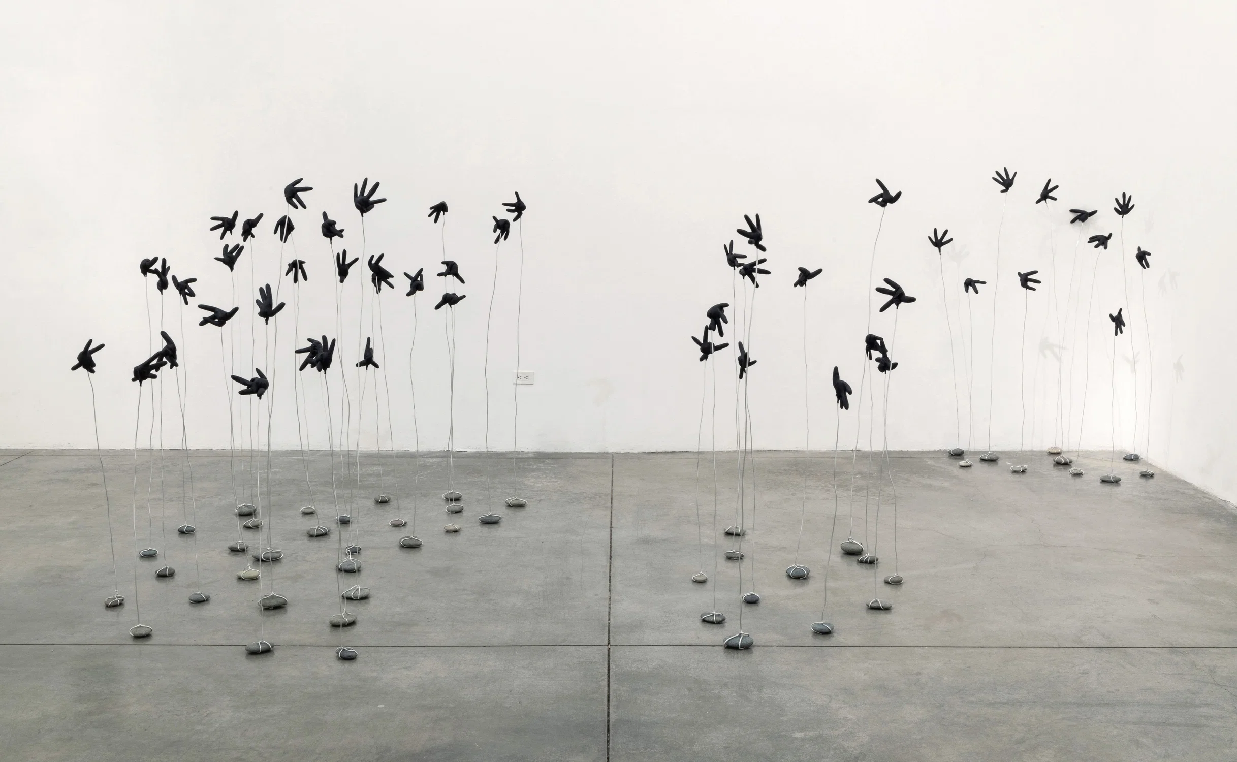 Dozens of pebbles on the ground, each wrapped in silver metal wire extending upwards, with the tops shaped like black gloves formed into flowers or flying birds.