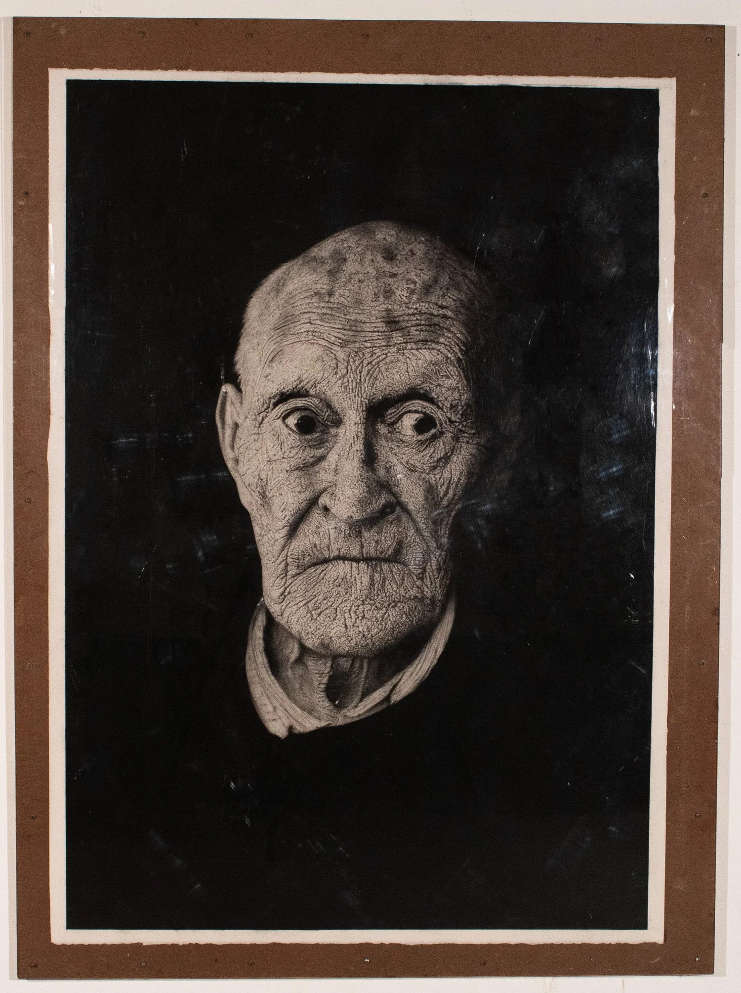 Charcoal portrait of an elderly man’s face, rendered in high contrast against a deep black background. The drawing emphasizes deeply etched wrinkles across his entire face with softened shading around the contours of his nose and eyes. His expression appears solemn and reflective, with fine charcoal markings capturing the texture of aged skin and the depth of his features emerging from the darkness.
